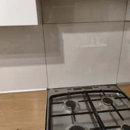 Błyszcząca, beżowa ściana nad kuchenką gazową, dopasowana kolorystycznie do szafek kuchennych, z widocznymi fugami i drobnymi zabrudzeniami na płycie gazowej.