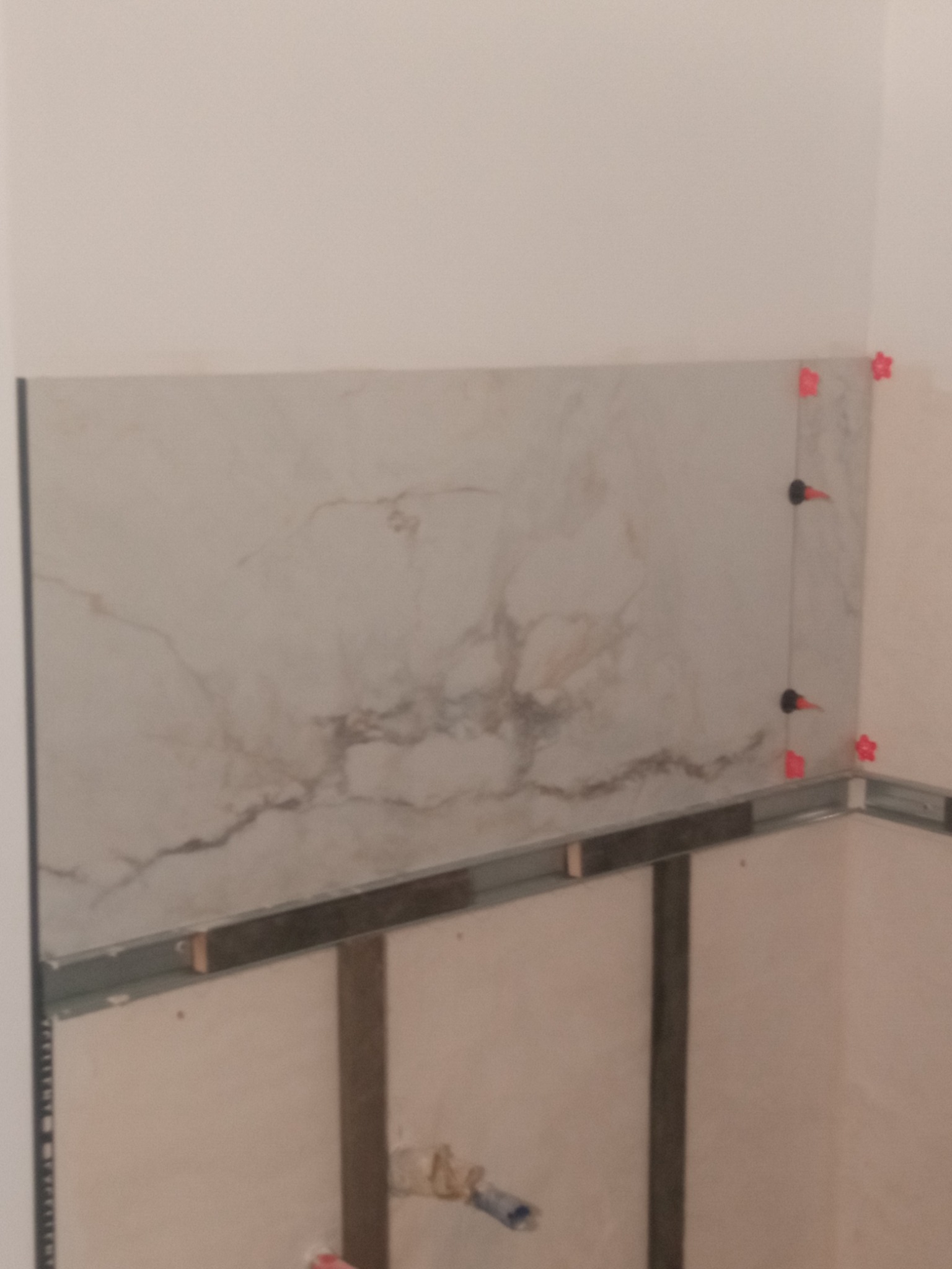 Fragment ściany w trakcie montażu płytek imitujących marmur, z użyciem poziomujących klipsów i listew, widoczne metalowe profile konstrukcyjne.