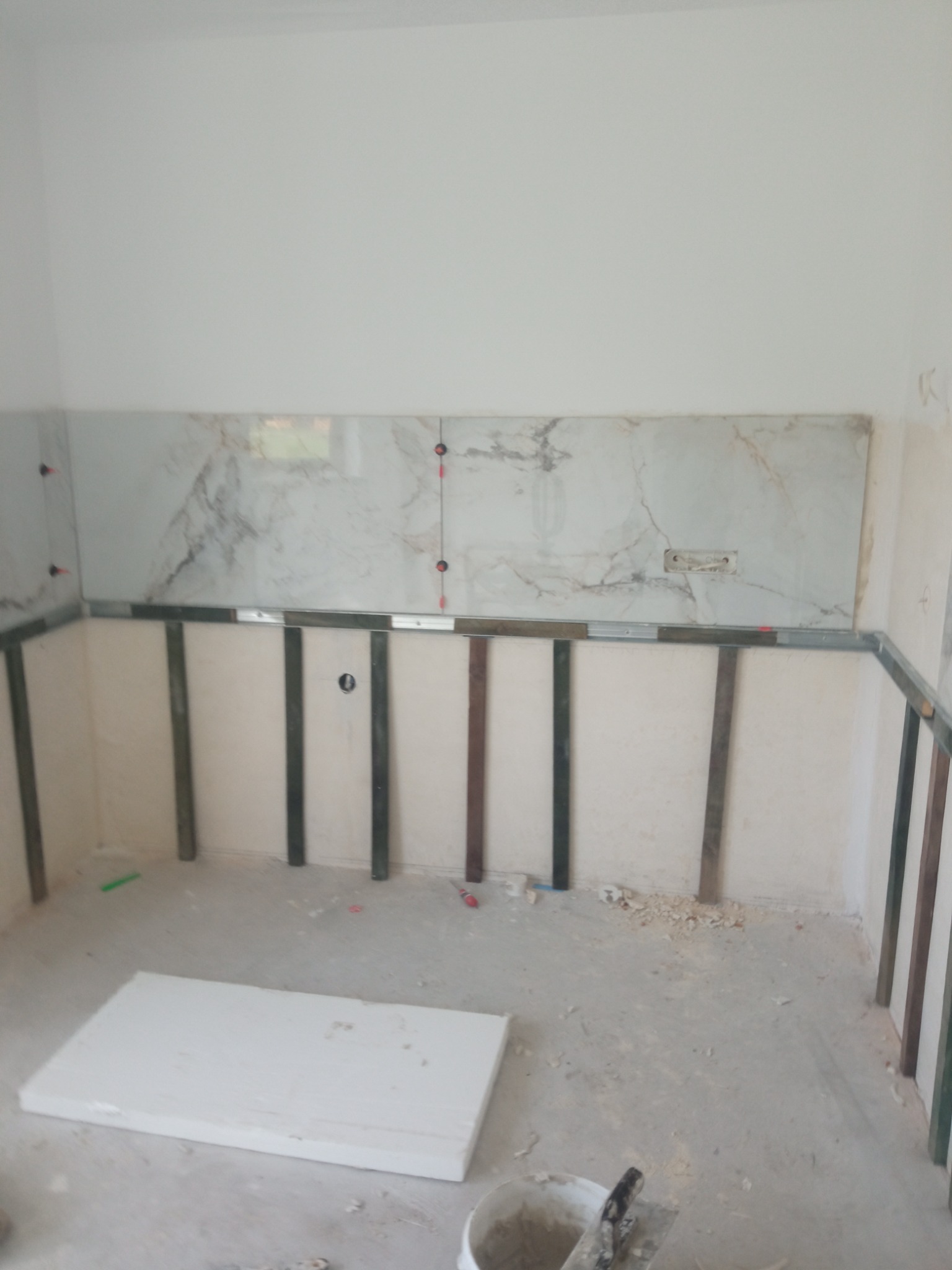 Montaż płytek imitujących marmur na ścianie, widoczne profile montażowe, poziomica laserowa oraz wiadro z zaprawą na podłodze.