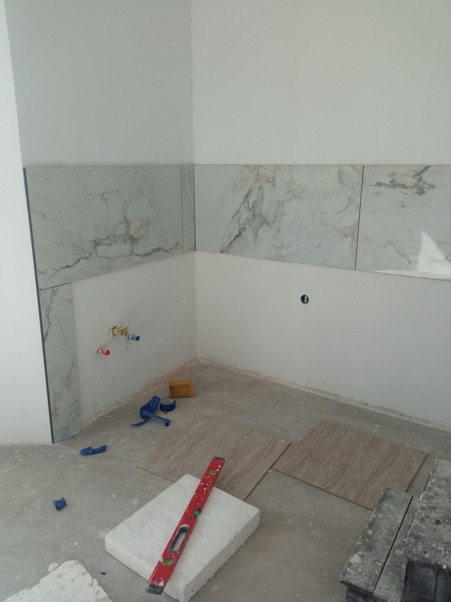 Fragment ściany z ułożonymi płytkami imitującymi marmur, widoczne wystające rury instalacji wodnej, poziomica na bloczku oraz płytki podłogowe przed fugowaniem.