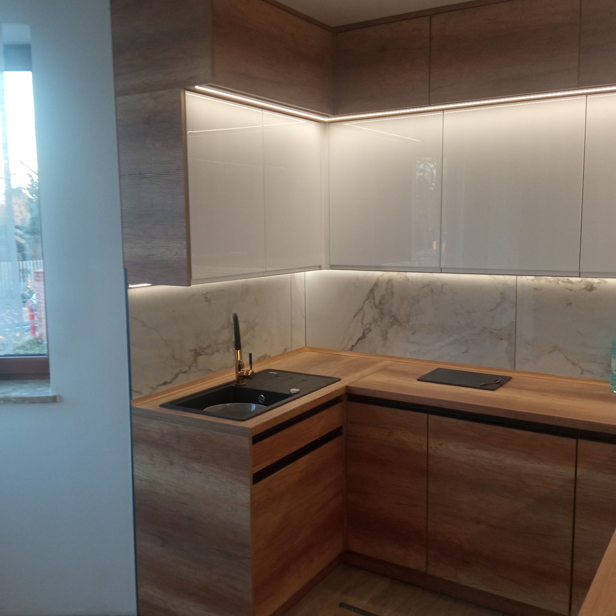 Nowoczesna kuchnia w odcieniach drewna i bieli, z marmurowym panelem nad blatem i oświetleniem LED pod szafkami wiszącymi, złota bateria przy czarnym zlewozmywaku.