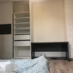 Wnętrze sypialni z widoczną białą szafą przesuwną z czarnym akcentem, biurkiem pod ścianą i łóżkiem w pierwszym planie z pościelą w roślinny wzór.