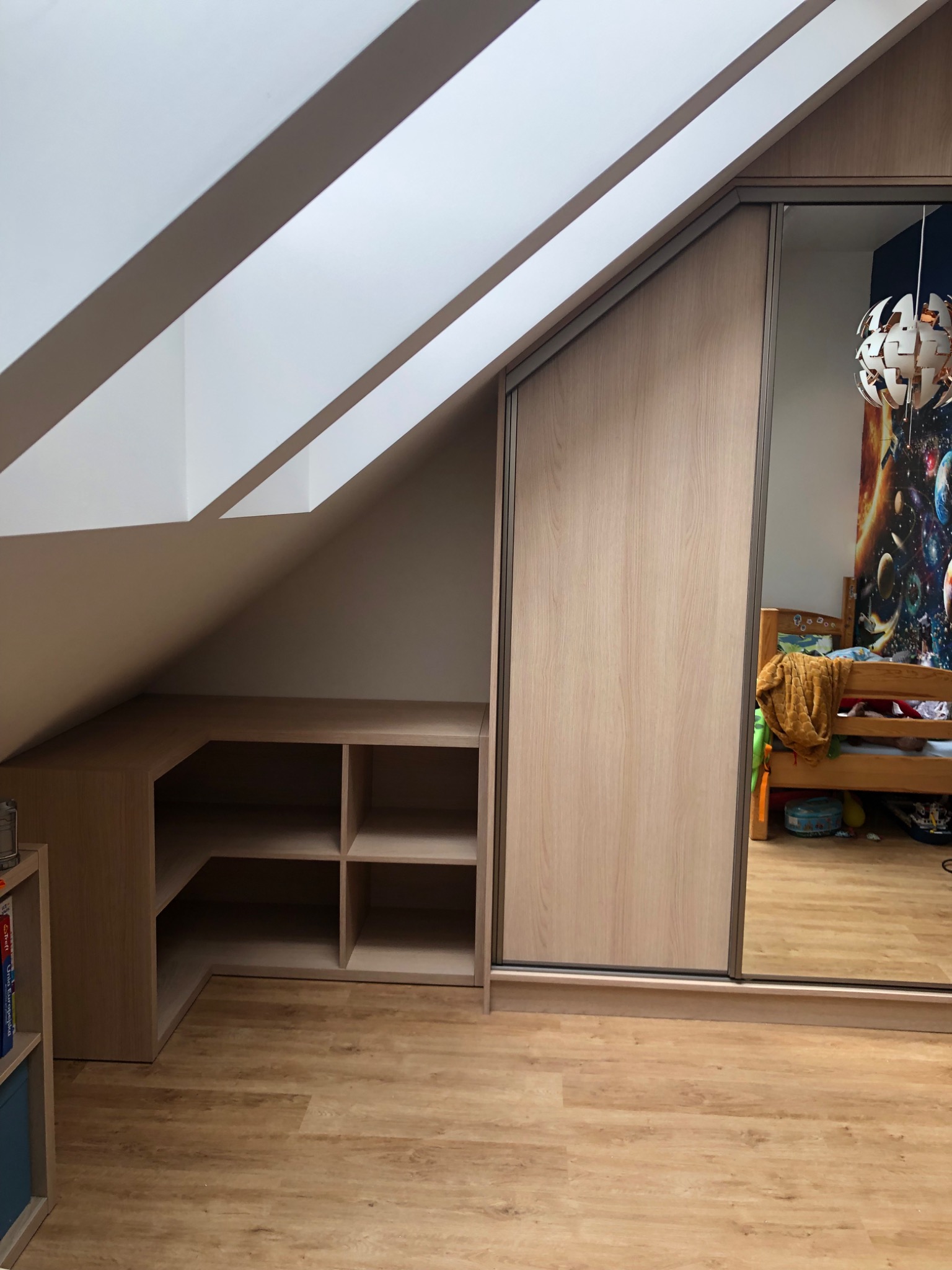 Zabudowa pod skosem na poddaszu z szafą przesuwną z lustrem i półkami, drewnopodobne wykończenie, widoczny pokój dziecka w odbiciu lustra.