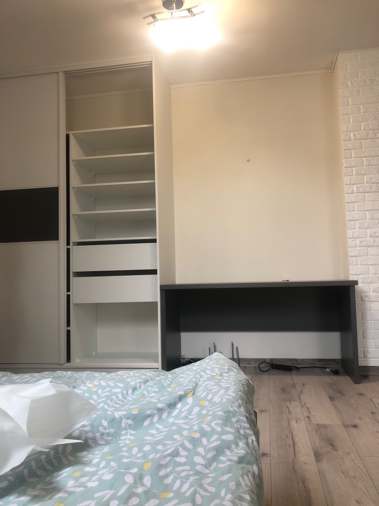 Wnętrze sypialni z widoczną białą szafą przesuwną z czarnym akcentem, biurkiem pod ścianą i łóżkiem w pierwszym planie z pościelą w roślinny wzór.