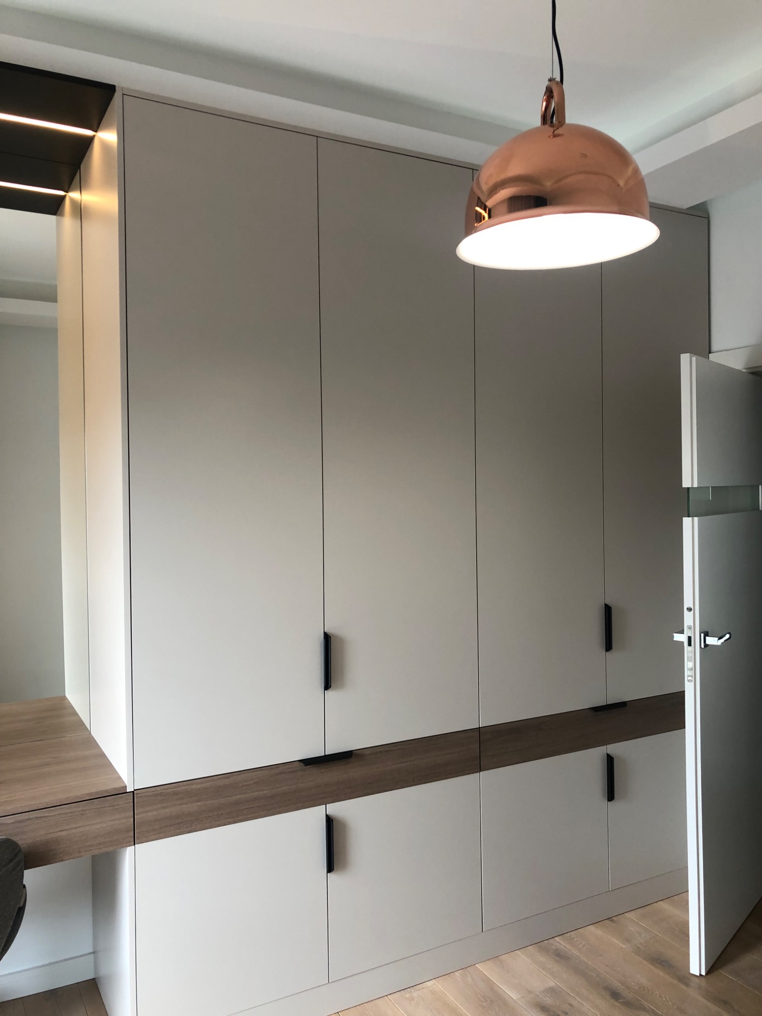 Wbudowana szafa z szarymi frontami i drewnianymi akcentami, zintegrowana z biurkiem, oświetlona miedzianą lampą wiszącą i listwami LED.