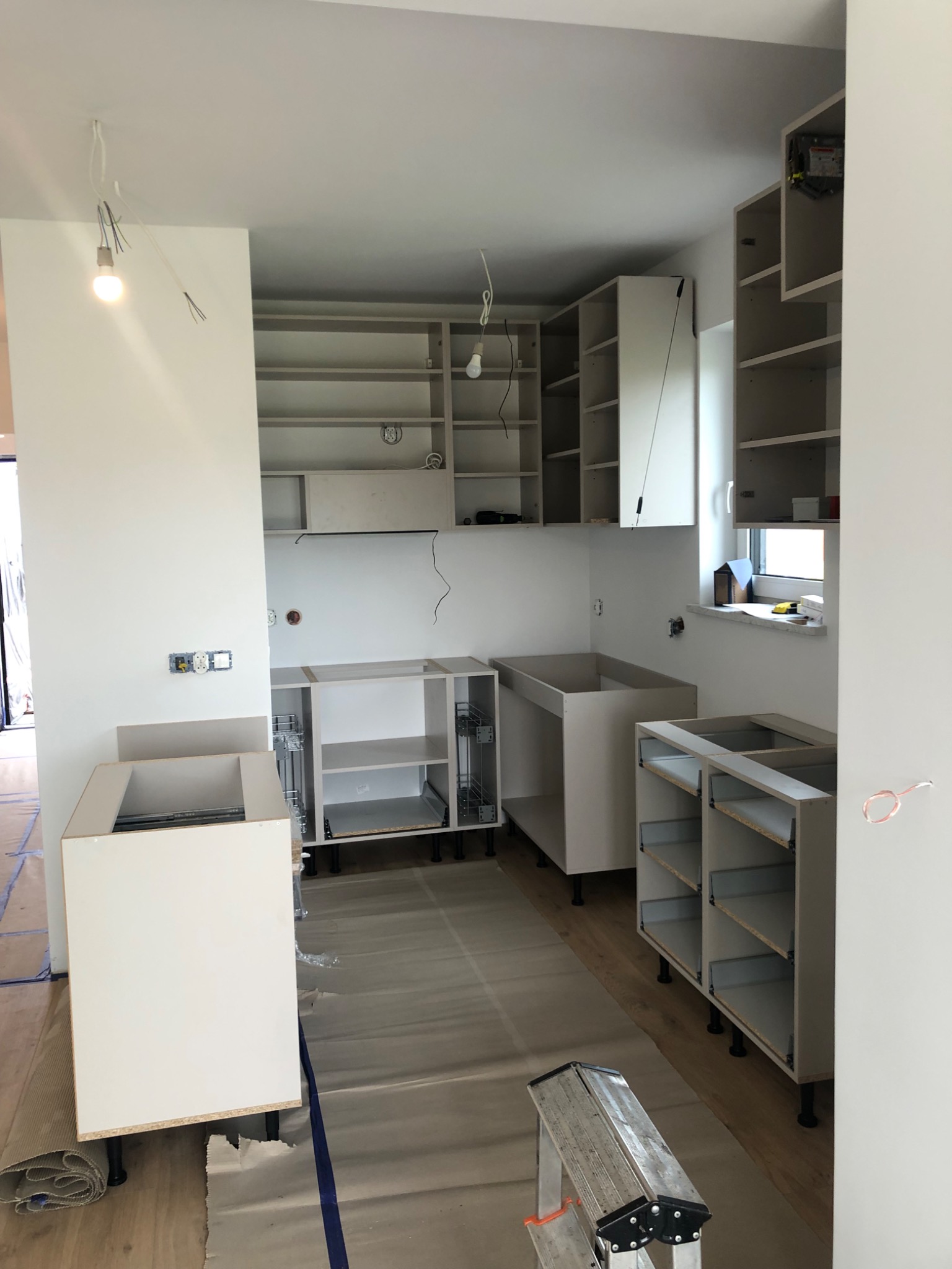 Wnętrze kuchni w trakcie montażu mebli: widoczne niedokończone szafki, drabina i zabezpieczona podłoga.