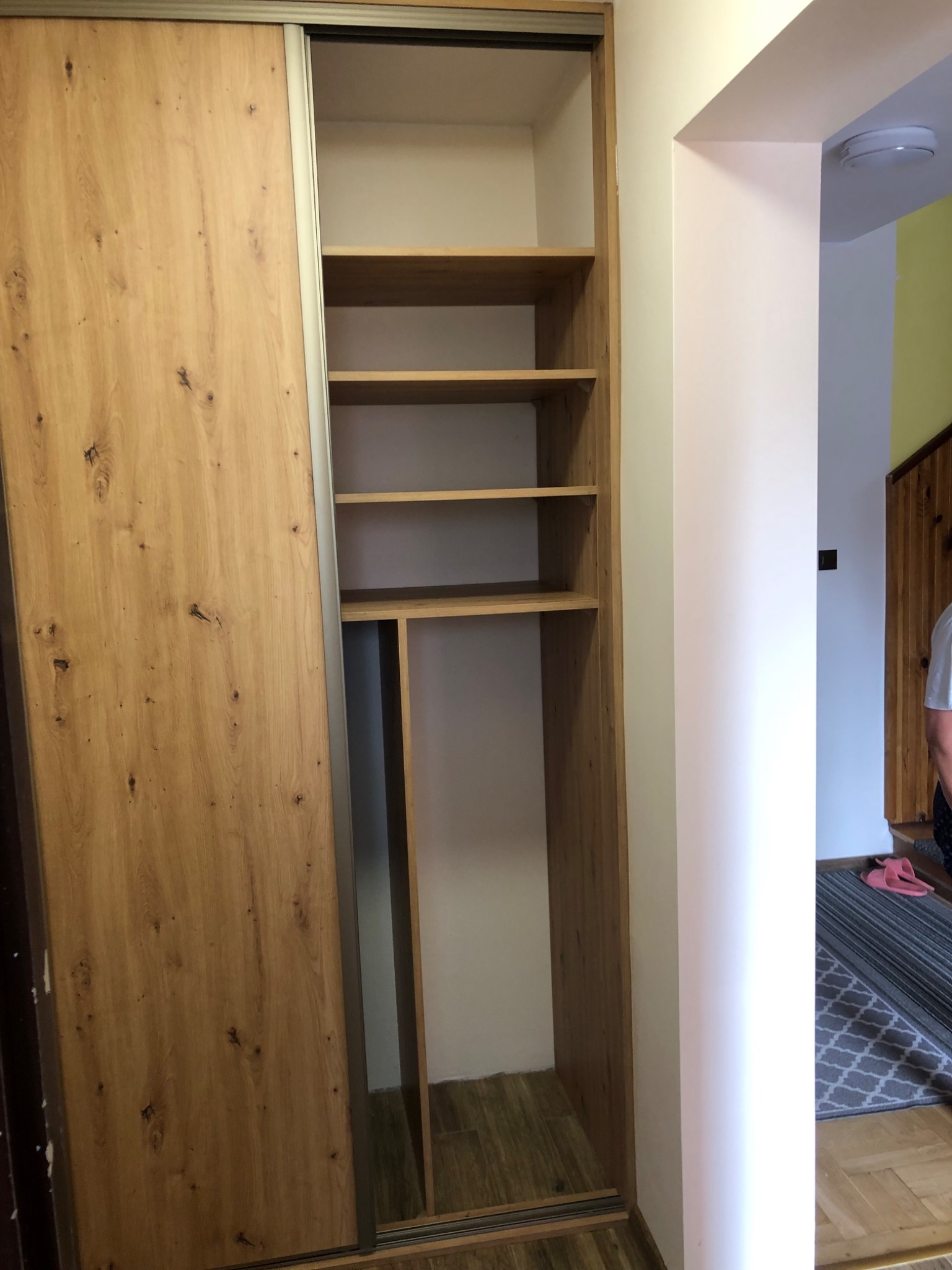 Wnętrze szafy wnękowej z przesuwnymi drzwiami o fakturze jasnego drewna, widoczne półki i drążek na ubrania, zarys wnętrza domu w tle.