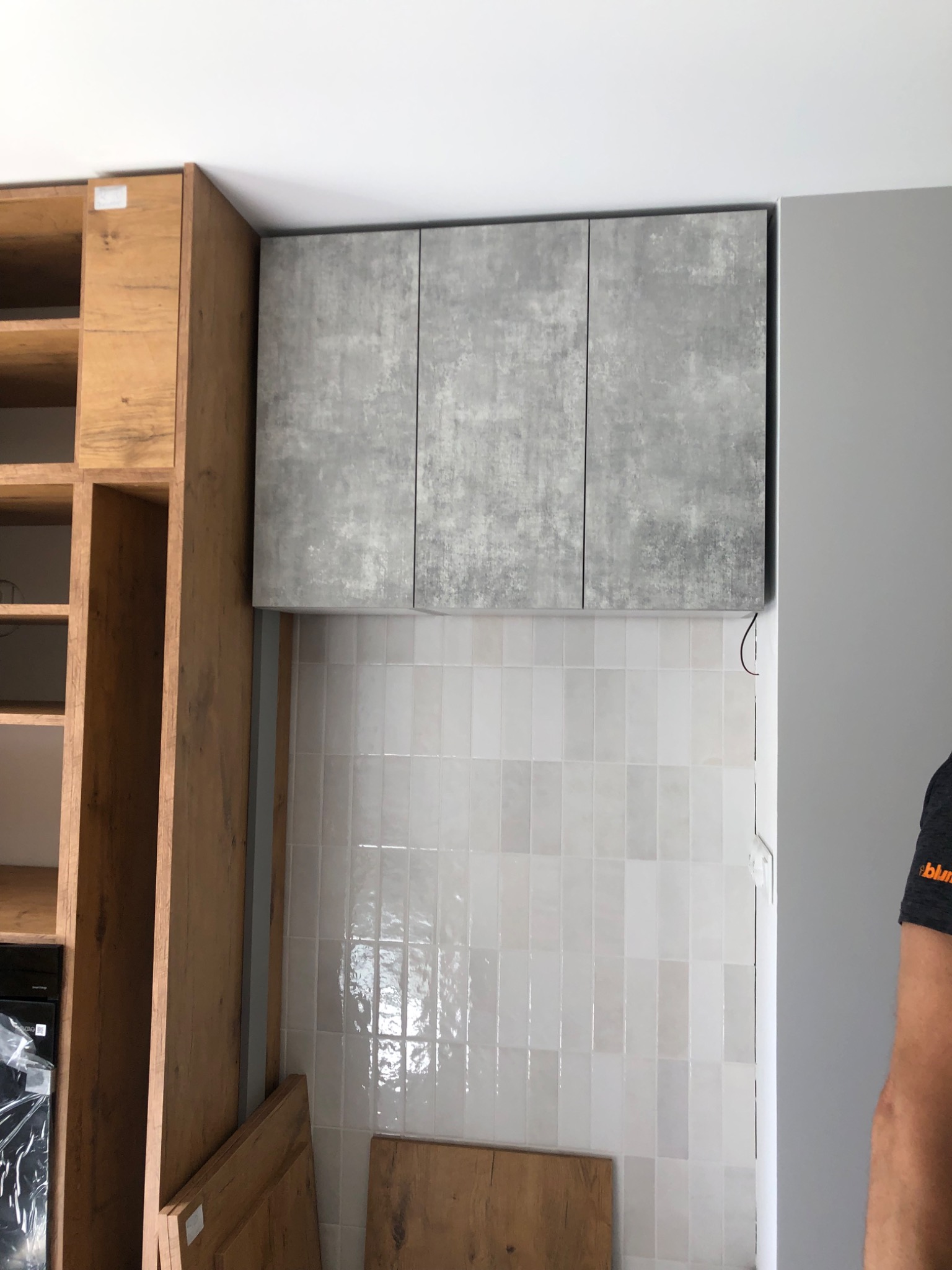 Częściowo zamontowana zabudowa kuchenna: drewniane szafki z otwartymi półkami obok szarych szafek wiszących o betonowej fakturze, białe płytki na ścianie.