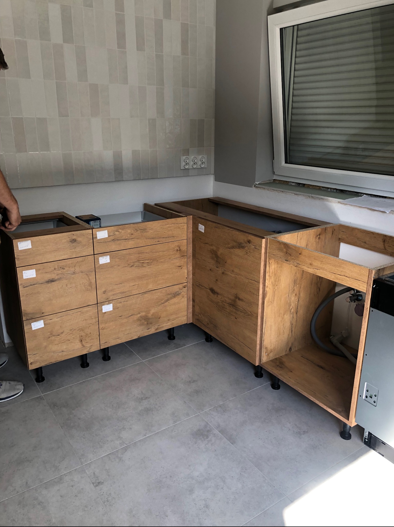 Częściowo zmontowana, drewniana zabudowa kuchenna z szufladami i szafkami, widoczna na tle szarych płytek i okna z roletą, z fragmentem dłoni montera.