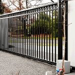 Czarna, metalowa brama przesuwna z pionowymi prętami, widoczny napęd elektryczny, szara nawierzchnia podjazdu, drzewa w tle.