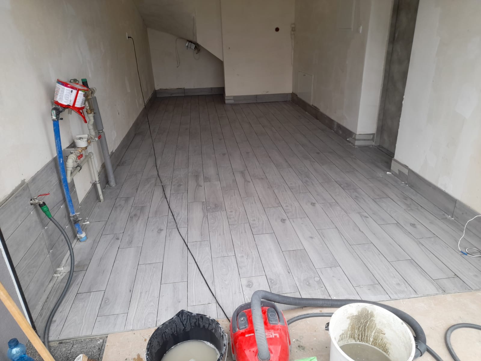 Ułożona podłoga z szarych płytek imitujących drewno w pomieszczeniu w trakcie prac wykończeniowych, widoczne narzędzia i materiały budowlane: odkurzacz, wiadro z zaprawą, węże doprowadzające wodę...