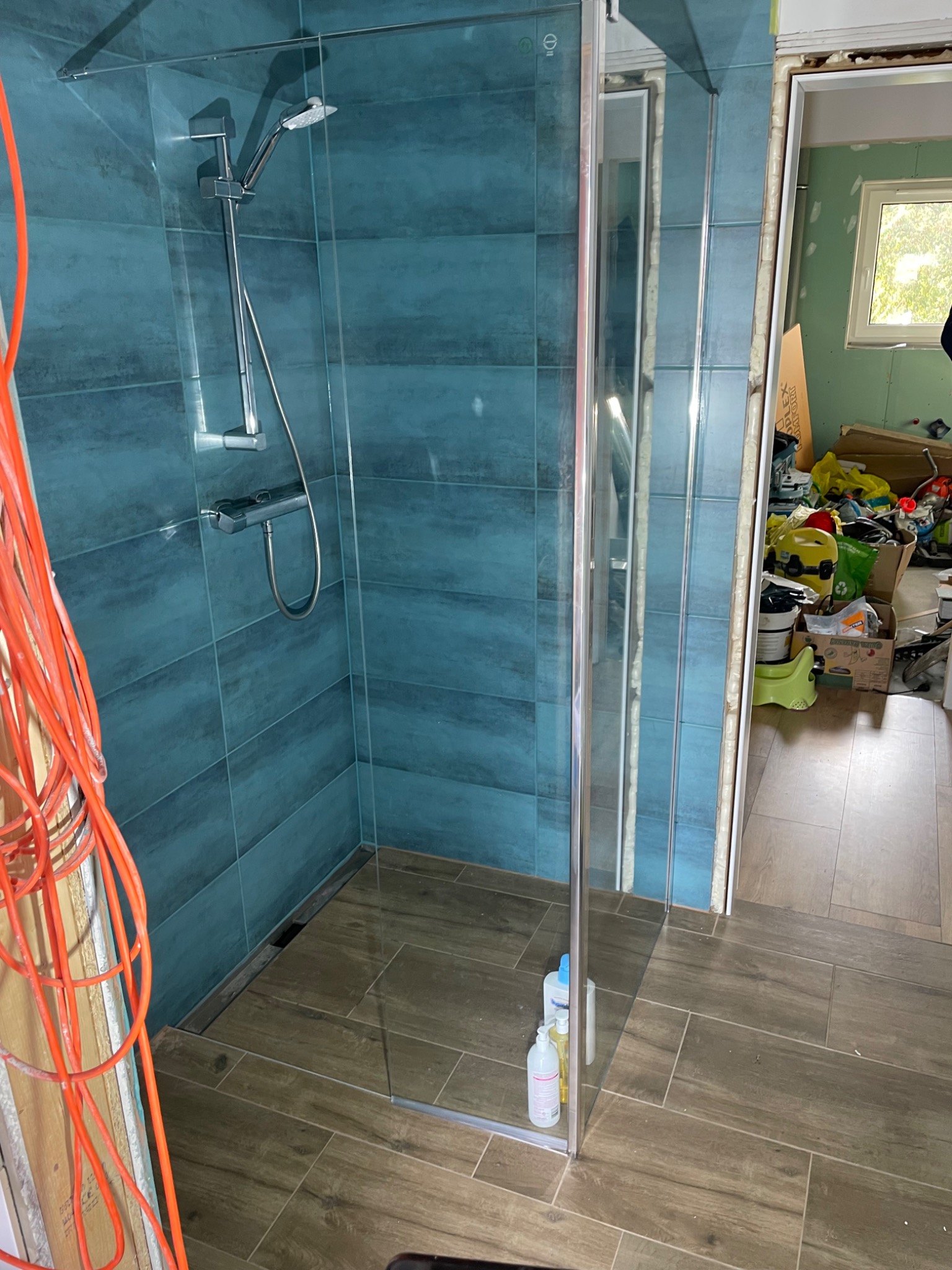 Nowoczesna kabina prysznicowa z turkusowymi płytkami i brązowymi płytkami podłogowymi, widoczne niedokończone prace wykończeniowe i pomarańczowe kable elektryczne.