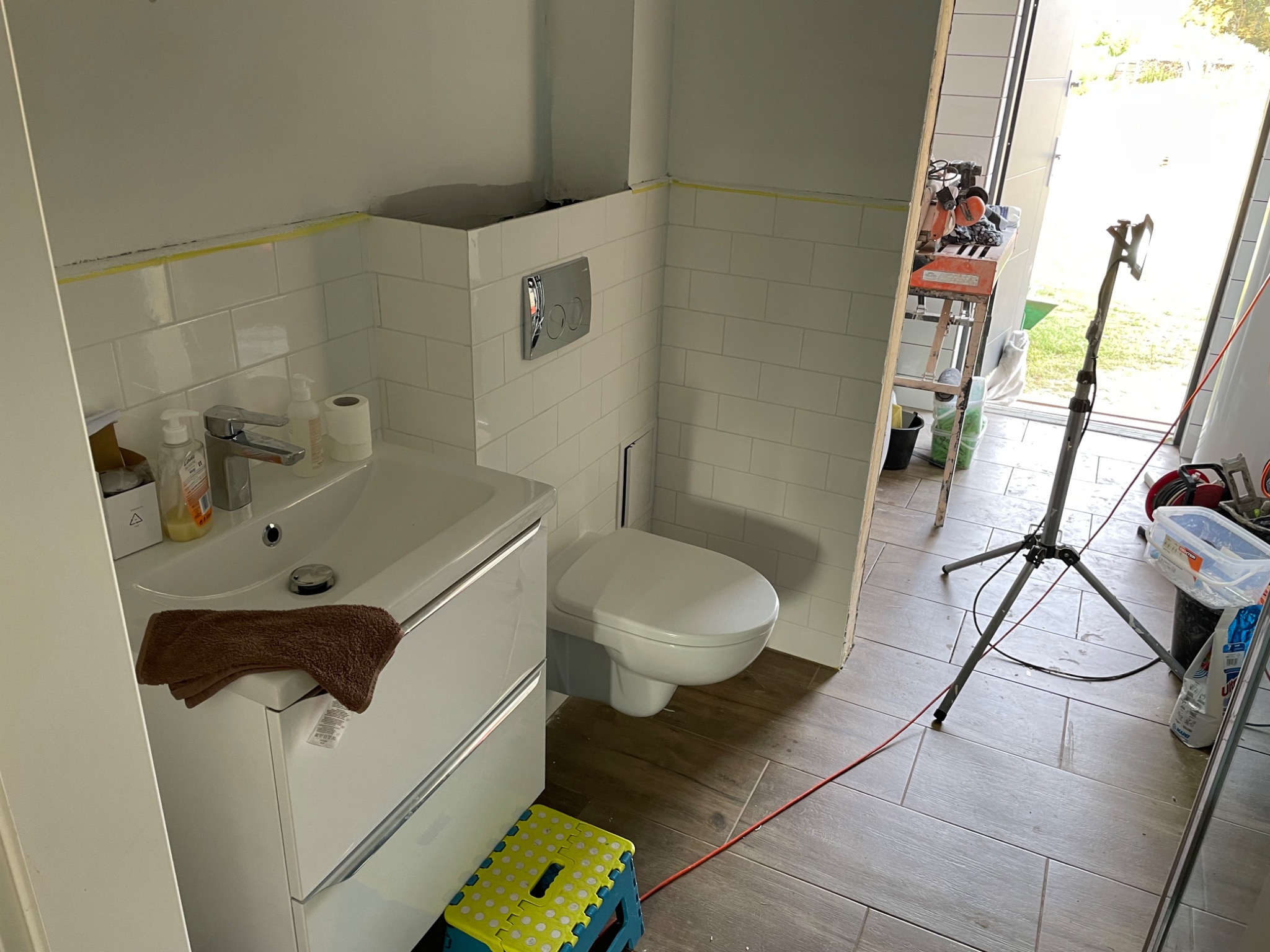 Wnętrze łazienki w trakcie prac, widoczna umywalka z szafką, podwieszana toaleta, białe płytki na ścianach z żółtą taśmą malarską, narzędzia glazurnicze i statyw oświetleniowy w tle, brązowy...