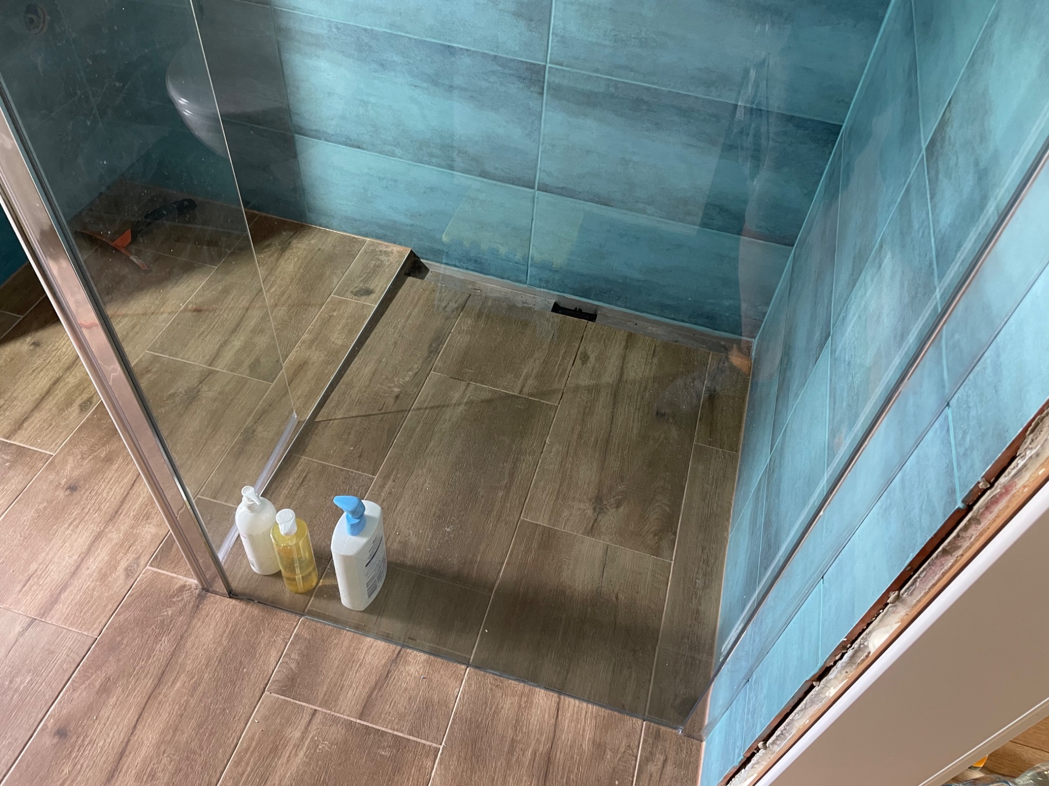 Kabina prysznicowa z niebieskimi płytkami na ścianach i brązowymi płytkami imitującymi drewno na podłodze, widoczne trzy butelki z kosmetykami w rogu kabiny.