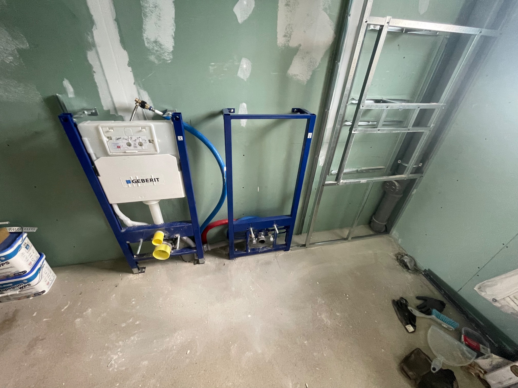 Stelaż podtynkowy Geberit zamontowany przy ścianie z płyt gipsowo-kartonowych w trakcie prac instalacyjnych, widoczne rury i elementy montażowe, obok stoją worki z materiałem budowlanym.