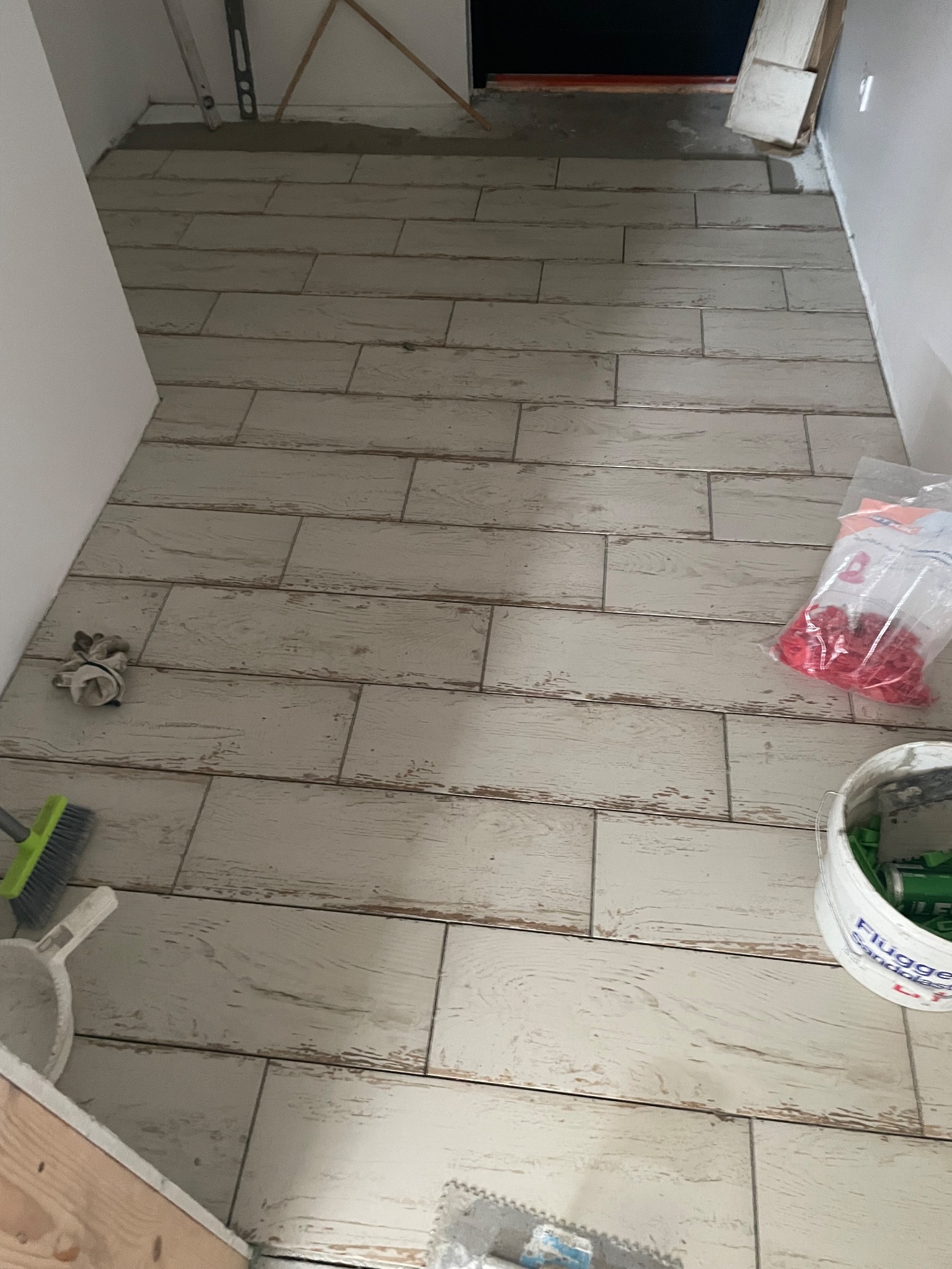 Ułożone podłogowe płytki imitujące drewno w łazience, widoczne narzędzia glazurnicze: poziomica, wiadro z klejem, paczka krzyżyków dystansowych.