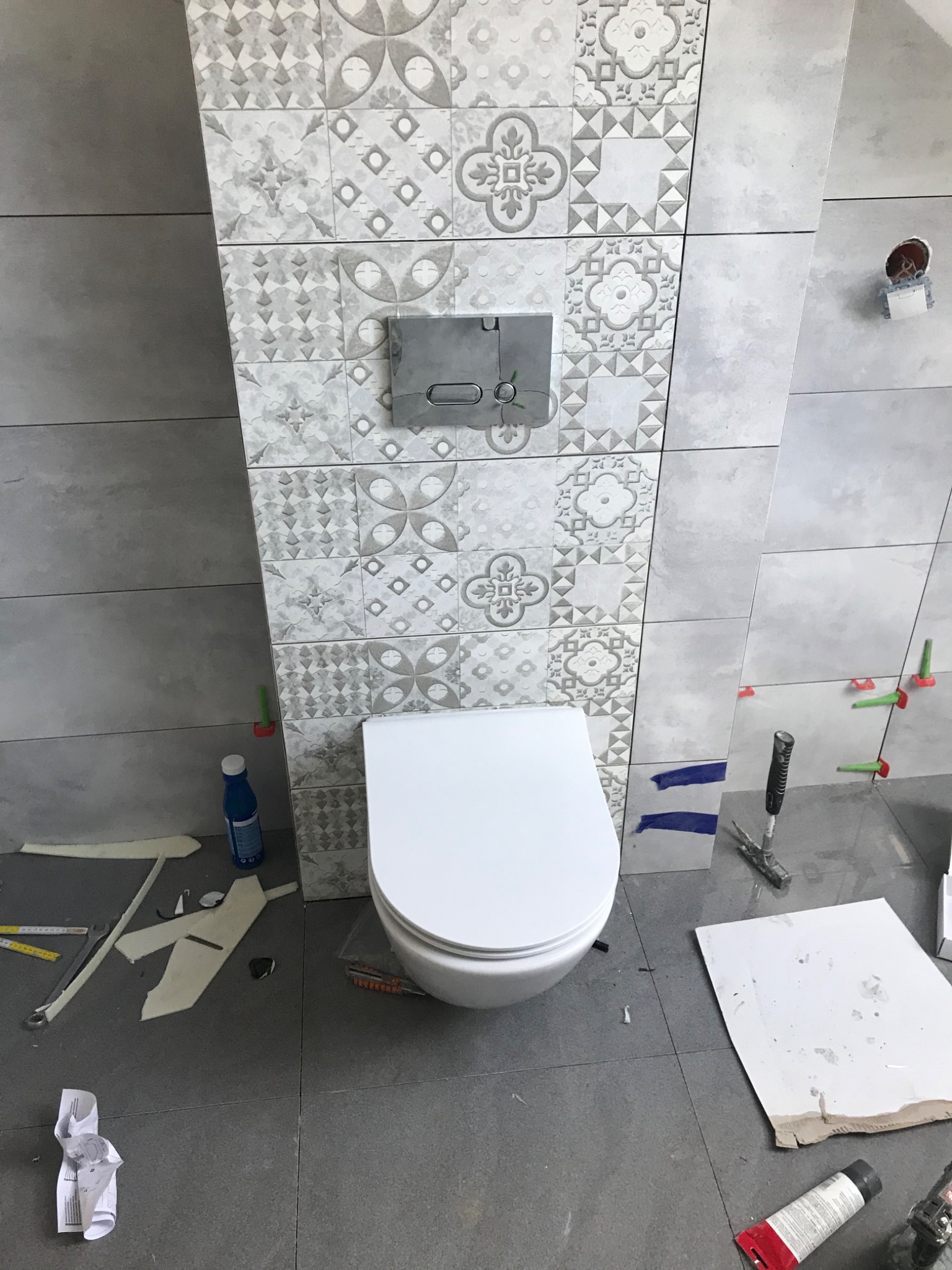 Montaż podtynkowego stelaża WC z ozdobnymi płytkami na ścianie, widoczne narzędzia i materiały budowlane na podłodze.