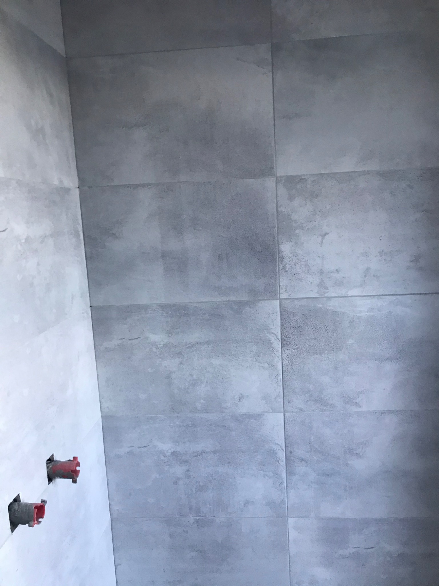 Ściana łazienki wyłożona szarymi płytkami imitującymi beton, z widocznymi otworami na instalację wodną z czerwonymi elementami.