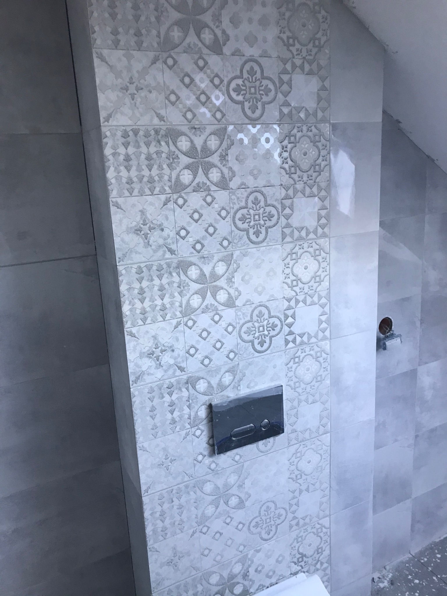 Ściana w łazience w Stargardzie wykończona dekoracyjnymi, jasnoszarymi płytkami ceramicznymi z motywami kwiatowymi i geometrycznymi. Widoczny przycisk spłuczki oraz fragment instalacji elektrycznej.