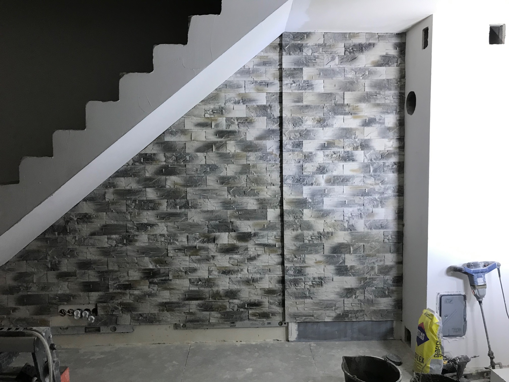 Ściana wyłożona szarą, nieregularną okładziną kamienną pod schodami, widoczne puszki elektryczne i narzędzia budowlane, worek zaprawy klejowej, w trakcie prac wykończeniowych.