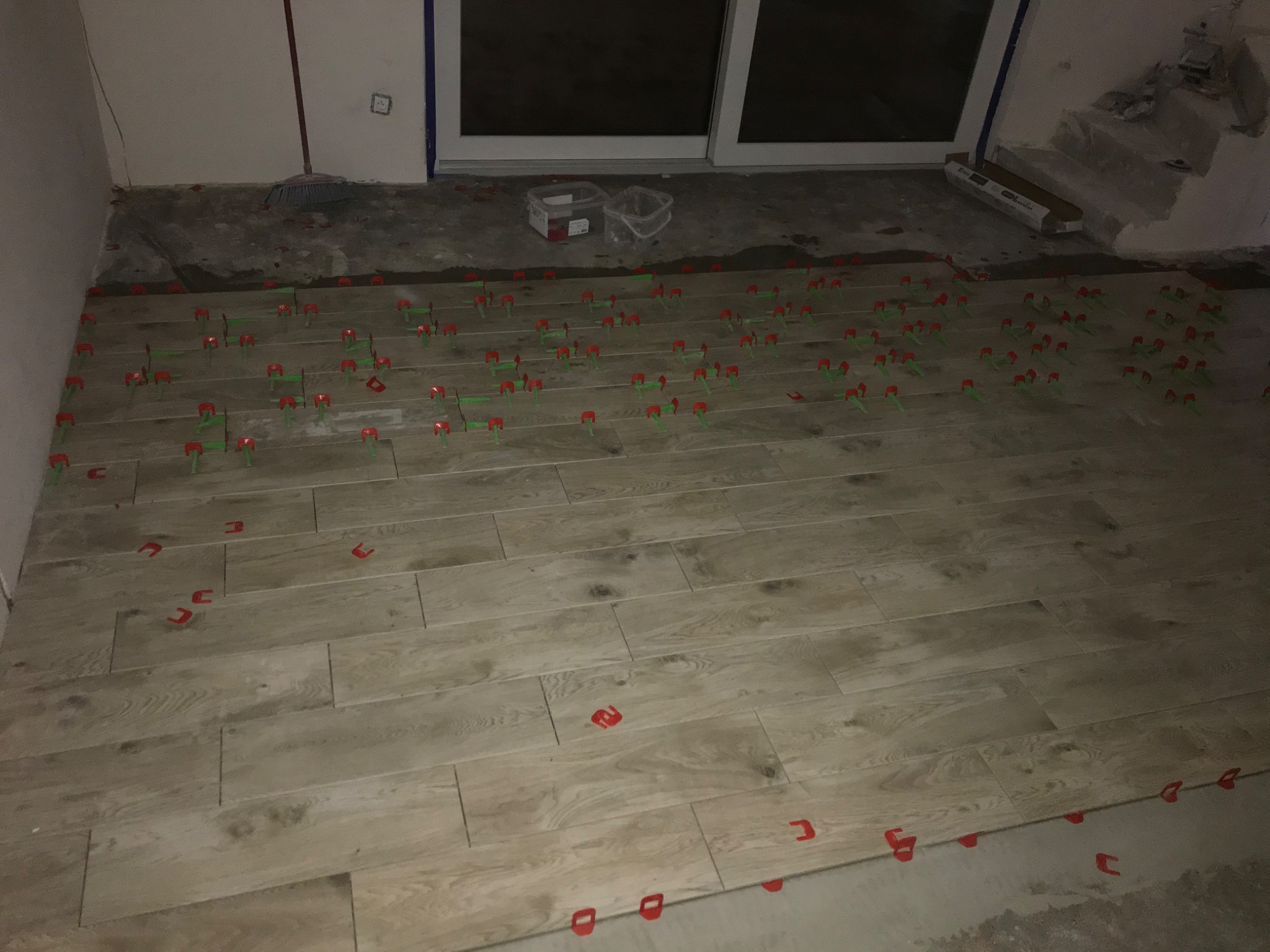 Układanie podłogi z płytek imitujących drewno w trakcie prac wykończeniowych, widoczne czerwone i zielone kliny poziomujące, szare fugi i fragmenty nieukończonej posadzki przy ścianach.