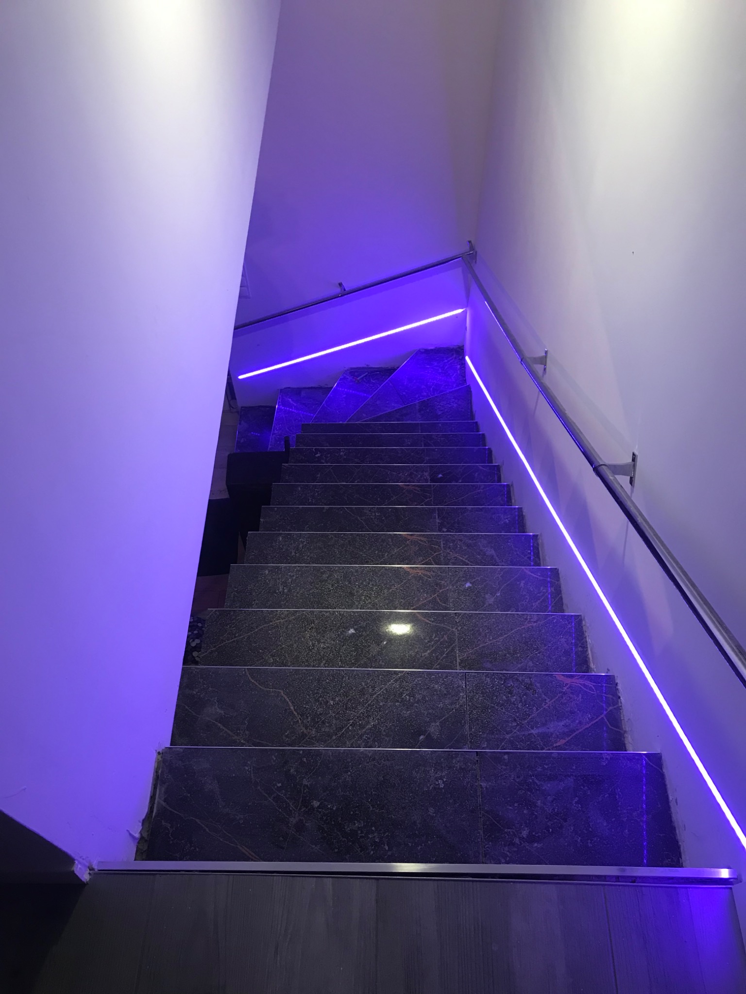 Schody wyłożone ciemnym gresem z marmurkowym wzorem, podświetlone neonowym, fioletowym światłem, widok z dołu.