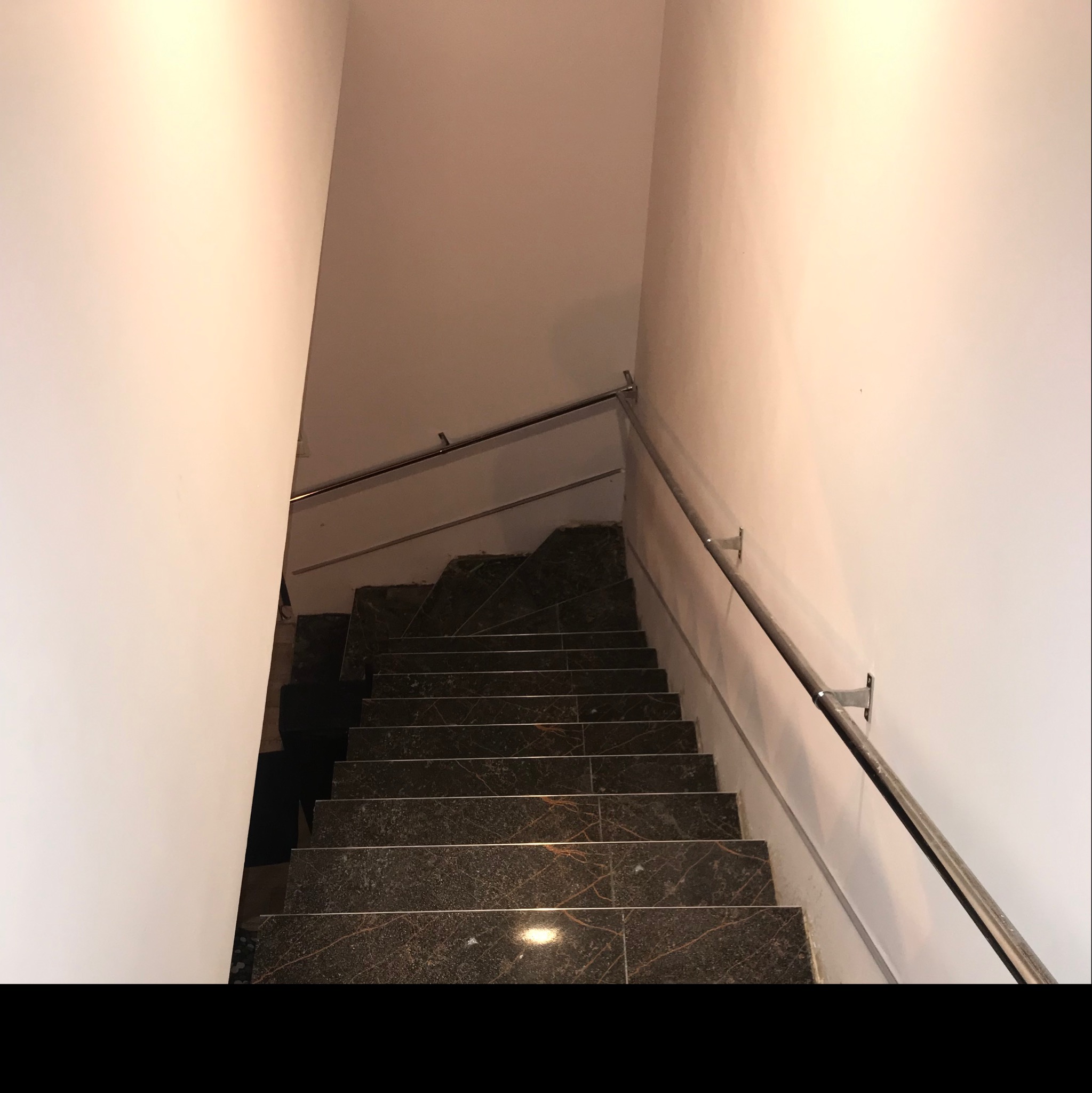 Schody wyłożone ciemnym gresem z wyraźnym, złotym żyłowaniem, prowadzące w dół, z zamontowaną metalową poręczą przy ścianie.