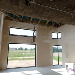 Firma usługowa Jacek Moś - Surowe ściany wewnętrzne w trakcie wykańczania, widoczne okna z czarnymi ramami i niedokończony sufit z odsłoniętą konstrukcją drewnianą i instalacjami.