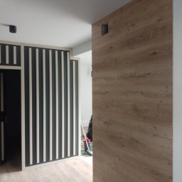 Firma usługowa Jacek Moś - Nowoczesne wykończenie wnętrza: ściana z paneli drewnopodobnych z ukrytym otworem wentylacyjnym, obok dekoracyjna ścianka działowa z pionowych listew w kolorze grafitowym i białym.