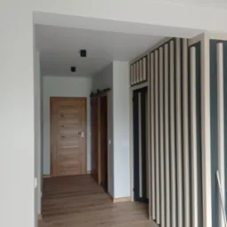 Wnętrze przedpokoju z drewnianą podłogą, dwoje drzwi, w tym jedne przesuwne na szynie, oraz dekoracyjna ścianka z pionowych listew w kolorach beżowym i granatowym.