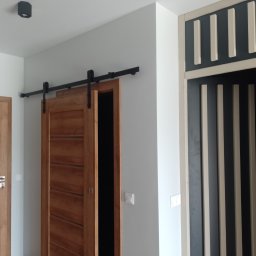 Firma usługowa Jacek Moś - Wnętrze z drewnianymi drzwiami przesuwnymi na czarnym szynoprzewodzie, widoczne fragmenty ściany w kolorze złamanej bieli, oraz dekoracyjne wykończenie ściany z pionowymi listwami w odcieniach...