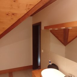 Firma usługowa Jacek Moś - Wykończona łazienka na poddaszu z drewnianą boazerią na suficie i ścianach, owalną białą umywalką na drewnianej szafce i dużym lustrem.