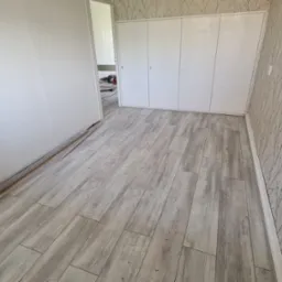 Jasne panele podłogowe imitujące drewno ułożone w pokoju z białą ścianą i tapetą w delikatny wzór gałązek. Przy ścianie leżą listwy przypodłogowe.