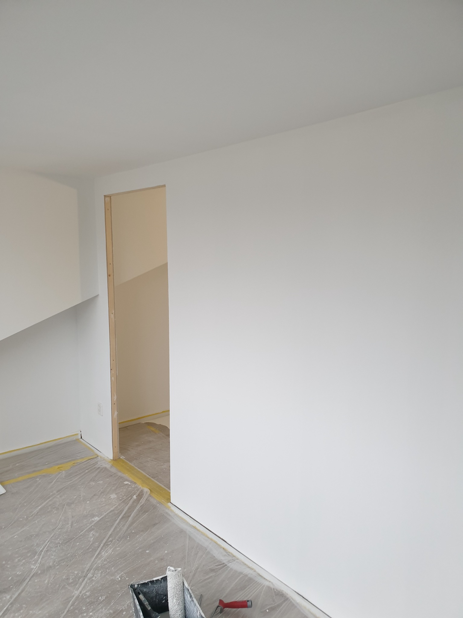 Świeżo pomalowane na biało ściany i sufit w pokoju, widoczne zabezpieczenia folią malarską i taśmą na podłodze oraz wiadro z farbą i wałek malarski w rogu pomieszczenia.