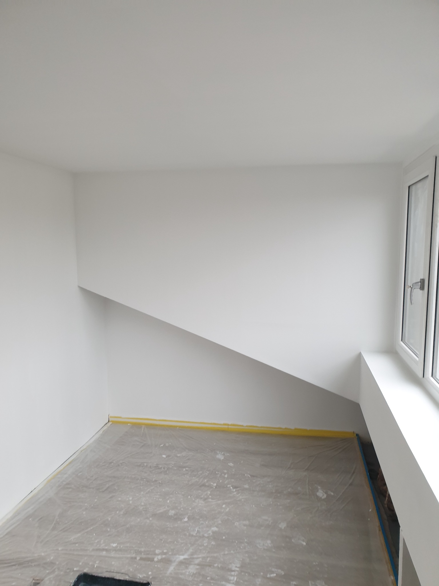 Pokój po świeżym pomalowaniu na biało, zabezpieczona podłoga folią i taśmą malarską, widoczne okno z białymi ramami.