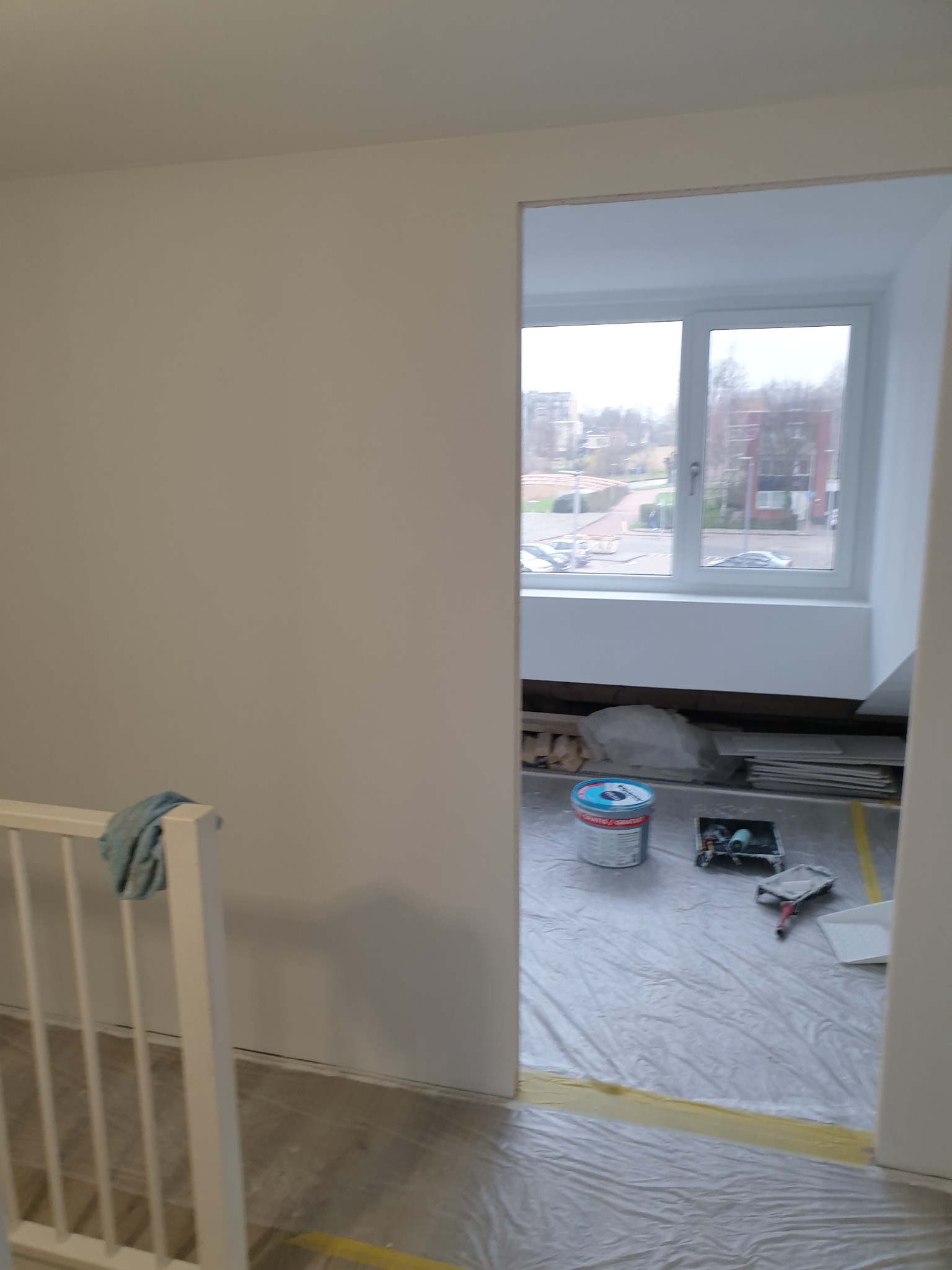 Świeżo pomalowane na biało ściany w pokoju, widoczny fragment balustrady, okno z widokiem na zabudowę miejską oraz wiadro z farbą i narzędzia malarskie zabezpieczone folią ochronną.