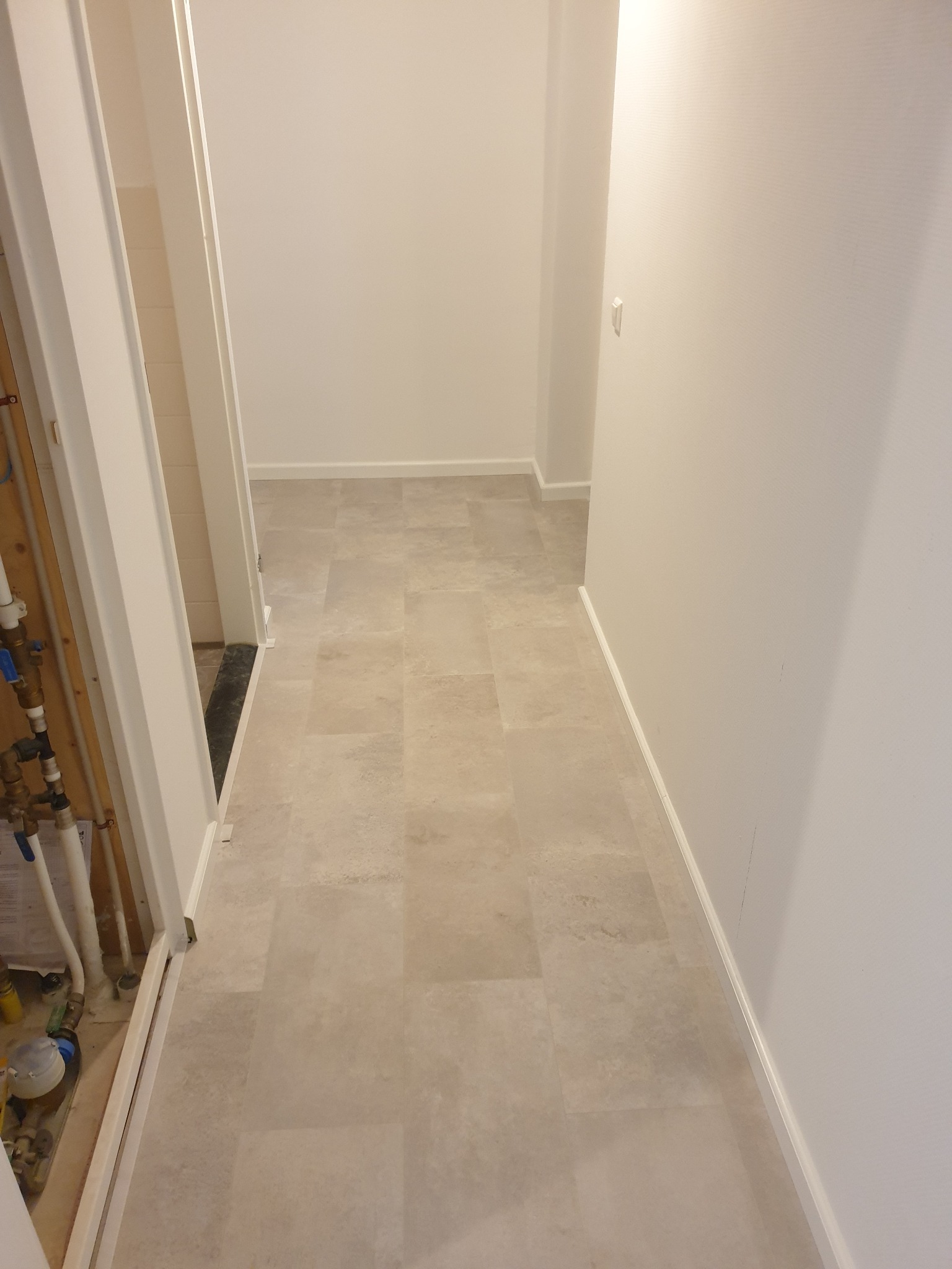 Jasna, świeżo ułożona podłoga z paneli w korytarzu, widoczne białe listwy przypodłogowe i fragment instalacji hydraulicznej w otwartym przejściu do pomieszczenia obok.