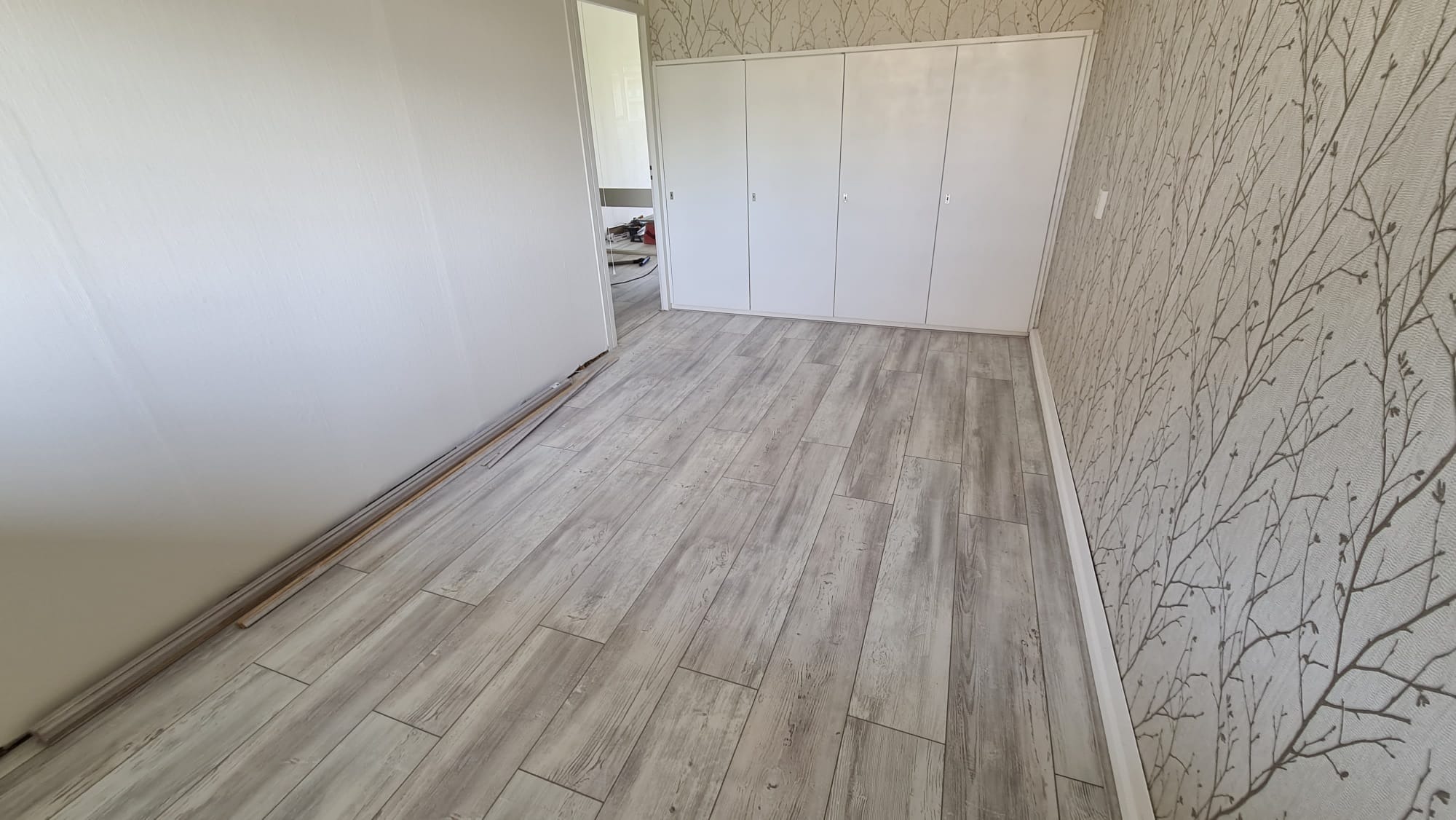 Jasne panele podłogowe imitujące drewno ułożone w pokoju z białą ścianą i tapetą w delikatny wzór gałązek. Przy ścianie leżą listwy przypodłogowe.