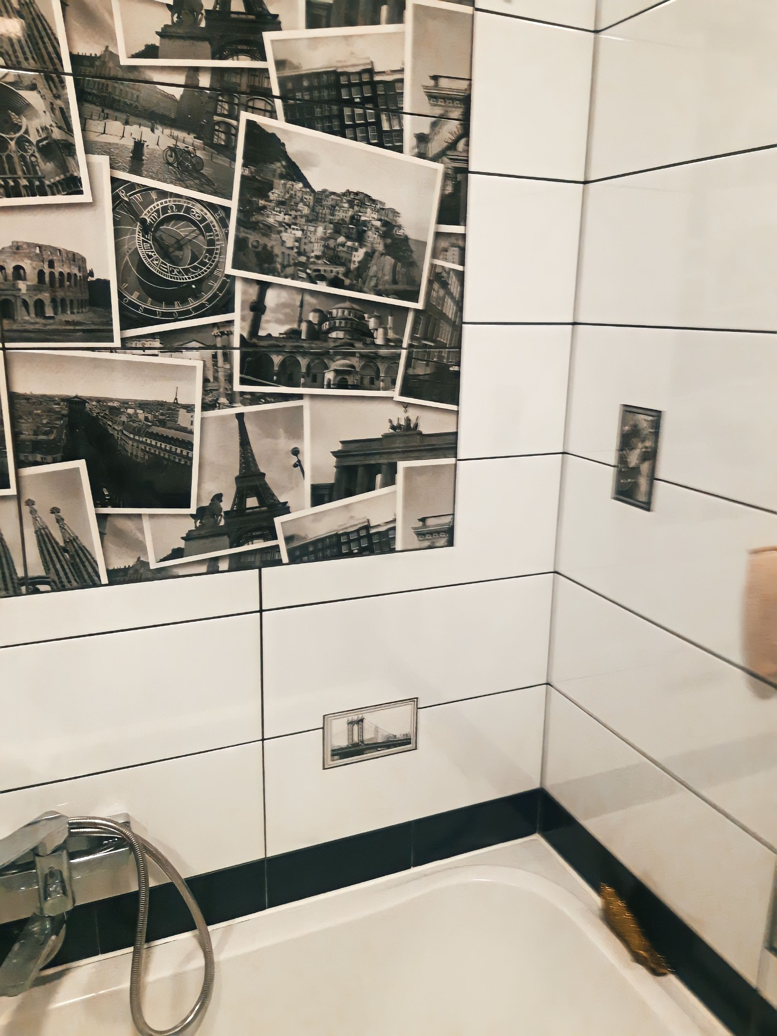 Białe płytki w łazience z dekoracyjnym motywem czarno-białych zdjęć zabytków i atrakcji turystycznych, widoczna bateria prysznicowa i fragment wanny.