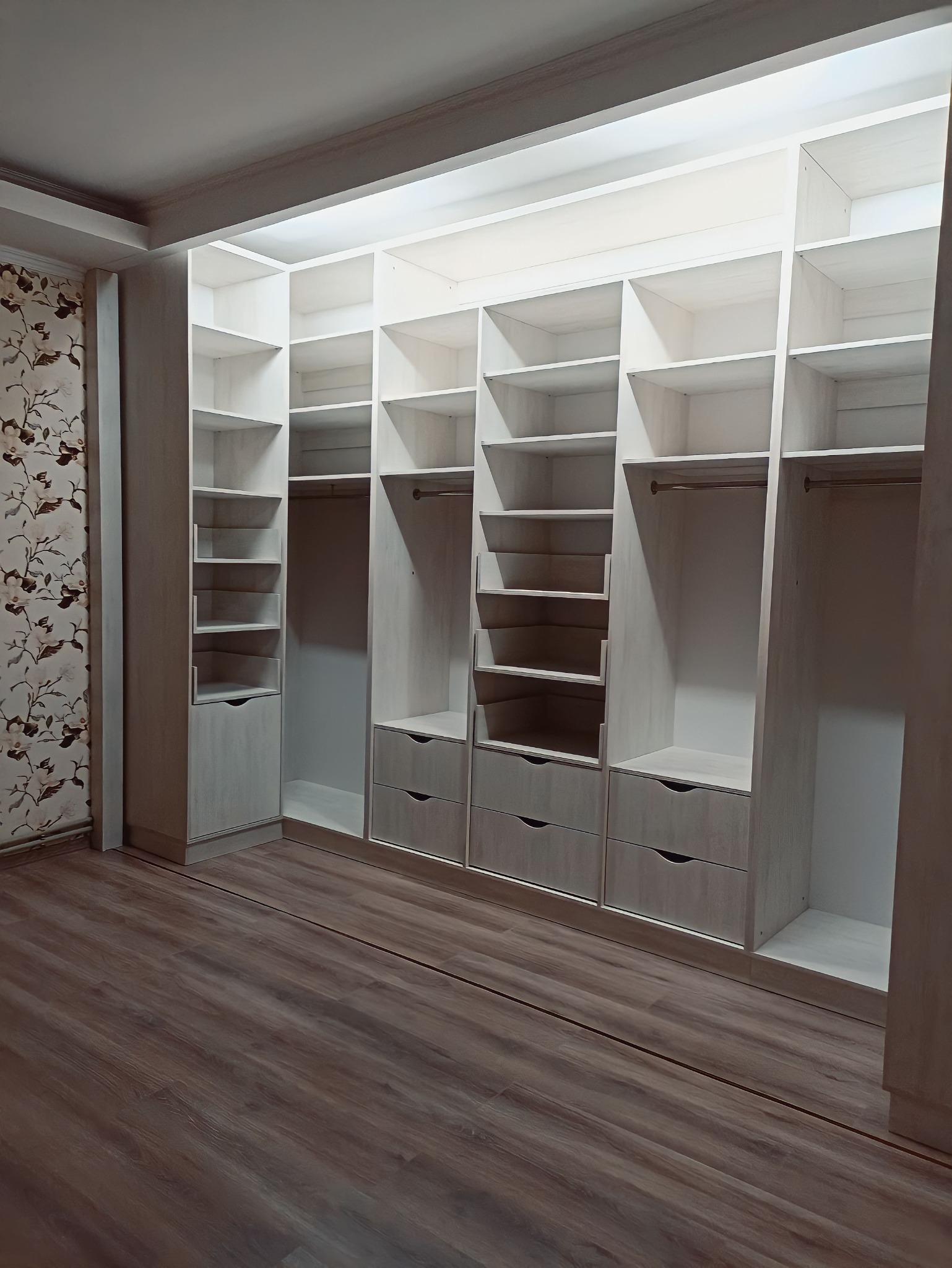 Wnętrze pokoju z zabudowaną, białą szafą na wymiar z szufladami i półkami, widoczne tapety w kwiaty na ścianie i panele podłogowe imitujące drewno.
