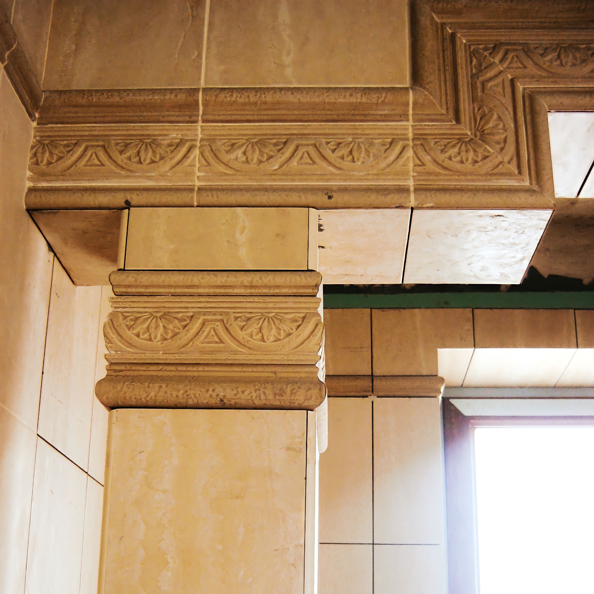 Fragment dekoracyjnego wykończenia wnętrza z beżowych płytek ceramicznych z ornamentami, widoczny fragment ściany i otworu okiennego.