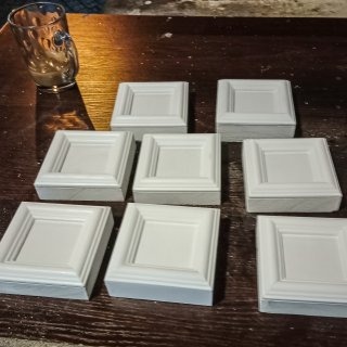 Siedem białych, kwadratowych paneli dekoracyjnych z wgłębieniem pośrodku, ułożonych na ciemnym, drewnianym blacie. W tle widoczna szklanka z płynem.