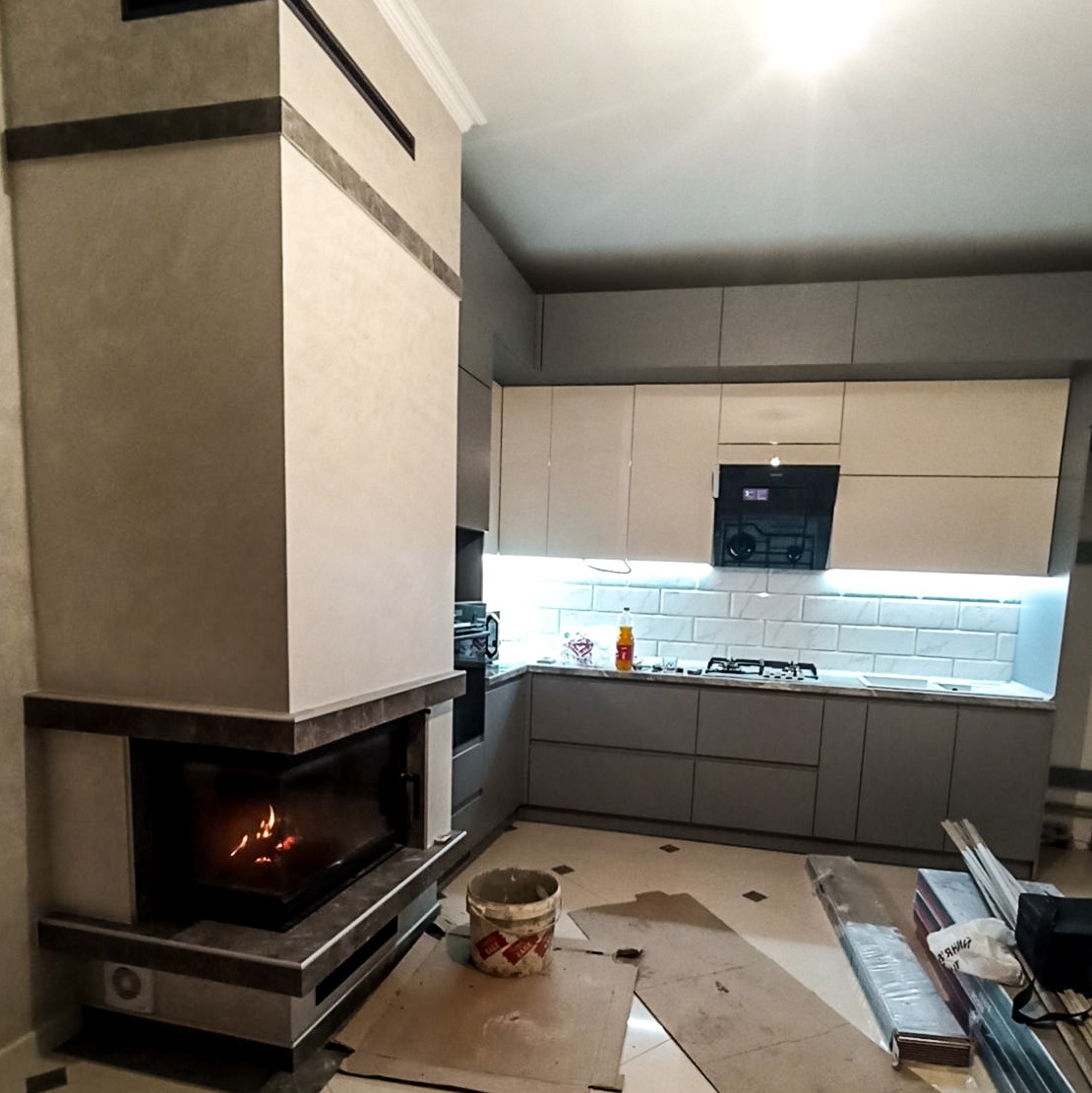 Nowoczesna kuchnia z kominkiem, szare szafki i białe płytki na ścianie, widoczne materiały budowlane na podłodze.