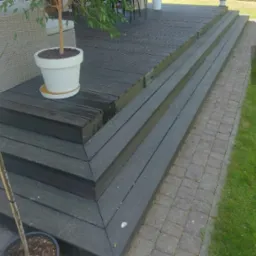 Ciemnoszara drewniana konstrukcja tarasu z kilkoma schodami, obok ścieżka z kostki brukowej i trawnik, w tle meble ogrodowe.