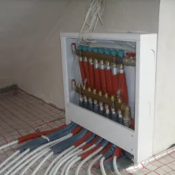 Rozdzielacz ogrzewania podłogowego w białej szafce, rury rozprowadzone na siatce montażowej, widoczne nieocieplone fragmenty rur oraz wystające przewody elektryczne.