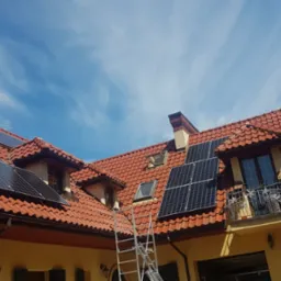 Montaż paneli fotowoltaicznych na dachu domu z czerwoną dachówką, widoczna drabina oparta o dach i wiatrowskaz w kształcie koguta.