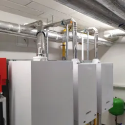 Trzy białe kotły grzewcze Viessmann Vitodens 200 z podłączeniami kominowymi i rurami w pomieszczeniu technicznym z izolowanymi rurami pod sufitem.