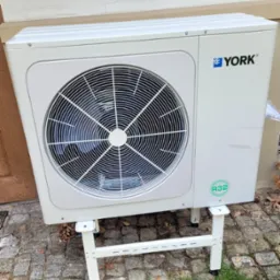 Biała jednostka zewnętrzna z wentylatorem marki YORK, umieszczona na metalowym stelażu, z widocznym oznaczeniem R32, ustawiona na brukowanym podłożu przy ścianie budynku.