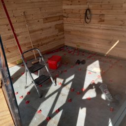 Kacper Dudra Stolarstwo-Montaż - Wnętrze drewnianego domku z częściowo ułożoną szarą podłogą z płytek, wykorzystującą czerwone klipsy poziomujące. Na podłodze widoczne narzędzia, wiadro i drabina. Ściany wyłożone boazerią.