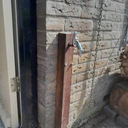 Kacper Dudra Stolarstwo-Montaż - Zewnętrzny kran zamontowany na drewnianym słupku przy ceglanej ścianie, obok drzwi z czarną ramą i wiszącym łańcuchem.